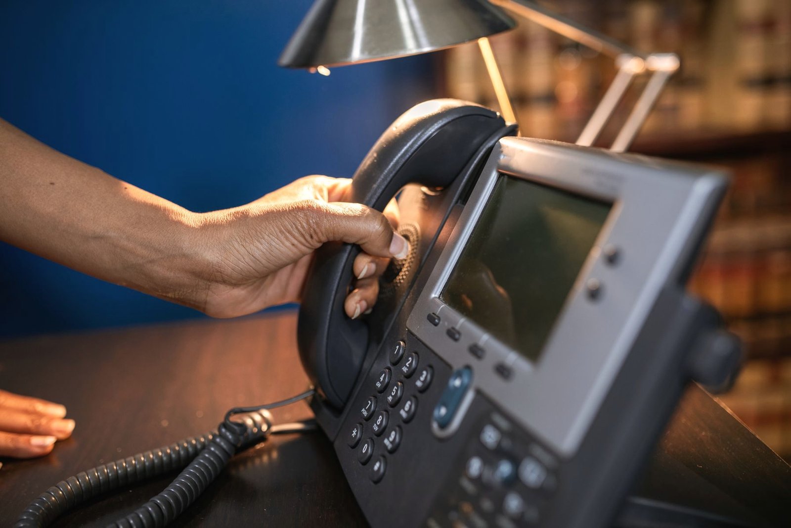 A hand lifts the handset from a desk phone under a reading lamp.