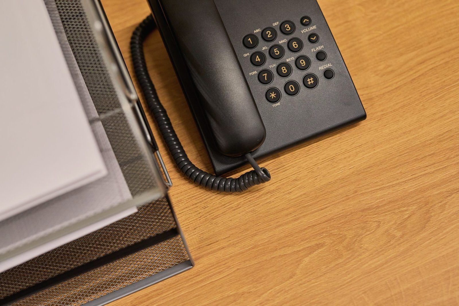 A black multi-line office telephone on a tidy wooden desk beside documents.