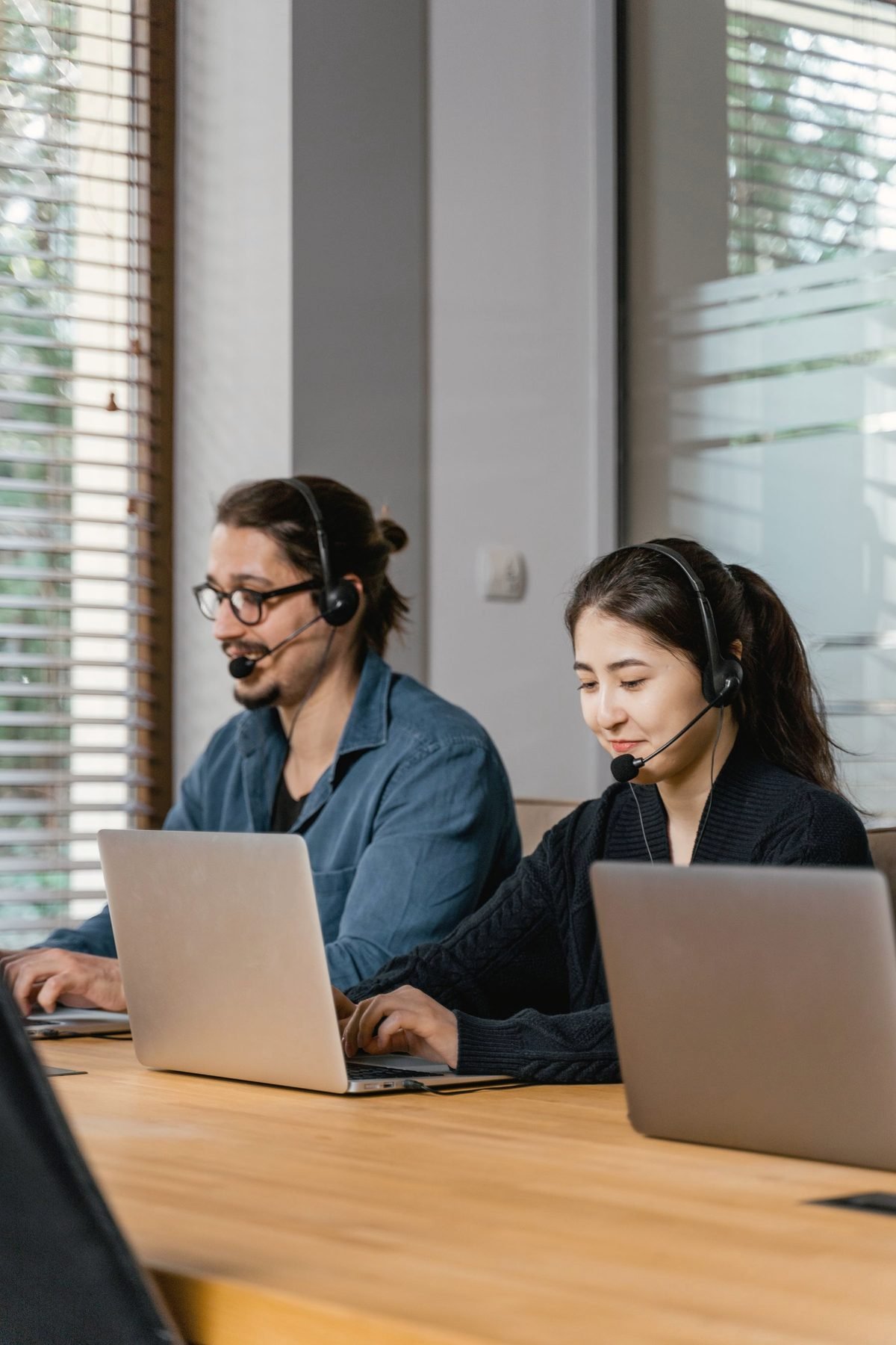 Two operators in headsets work at laptops across a table.